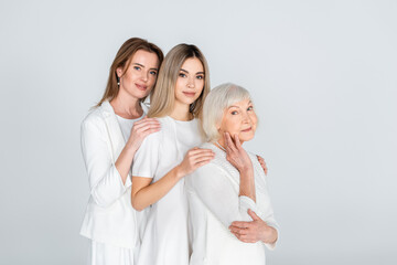 three generation of joyful women smiling while looking at camera and hugging isolated on grey