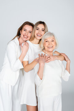 three generation of smiling women looking at camera and hugging isolated on grey