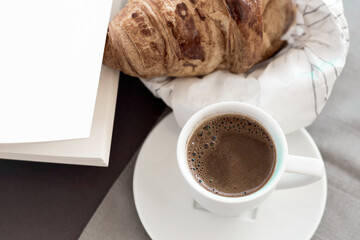 Obraz na płótnie Canvas Morning coffee concept, white cup of coffee, empty book page, fresh baked croussant in paper pack on gray cloth and dark grey background