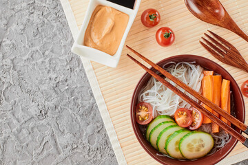 Fototapeta na wymiar Asian noodle soup, ramen with vegetables in a bowl with wooden chopsticks on a bamboo mat top view. Ingredients carrots, cucumbers, tomatoes, noodles.