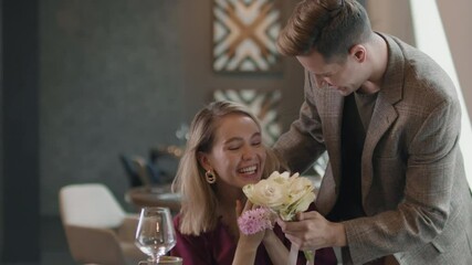 Medium shot of man closing eyes from behind to beautiful woman sitting in cafe with cup of coffee waiting for him having date