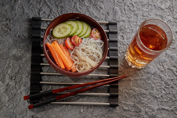Asian noodle soup, Ramen with vegetables in a bowl on a stand for hot dishes. Gray textured background top view. Ingredients carrots, cucumbers, tomatoes, noodles.