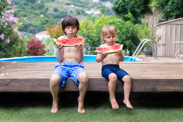 Two siblings brothers with big red pieces of watermelon sitting near the swimming pool  during summer holidays. Healthy food at vacances