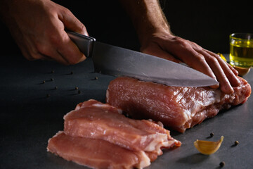 Cooking. Male hands with a knife cut a large piece of raw fresh meat on a stone gray background next to spices