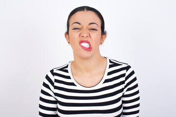 Young beautiful woman wearing stripped t-shirt against white background keeps teeth clenched, frowns face in dissatisfaction, irritated because of much duties.