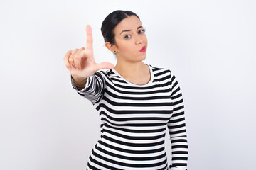 Young beautiful woman wearing stripped t-shirt against white background making fun of people with fingers on forehead doing loser gesture mocking and insulting.