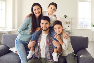 Happy mother, father and cute children on sofa in the living-room. Webcam portrait of smiling young couple with charming kids sitting on couch at home and looking at camera during family video call