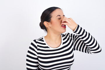 Young beautiful woman wearing stripped t-shirt against white background smelling something stinky and disgusting, intolerable smell, holding breath with fingers on nose. Bad smell