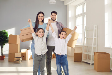 Fototapeta na wymiar Happy family with little children celebrating moving into new home. Portrait of smiling young couple with excited kids standing in room with packed boxes. Buying house, real estate, mortgage concept