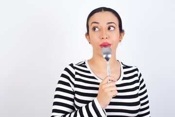 Very hungry Young beautiful woman wearing stripped t-shirt against white background holding spoon into mouth dream of tasty meal