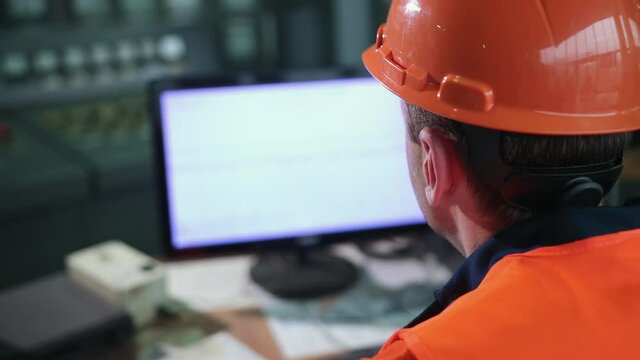 Inside heavy industry, factory male industrial engineer works on personal computer. Industrial engineer in hard hat wearing safety jacket uses computer. Works at heavy industry manufacturing factory