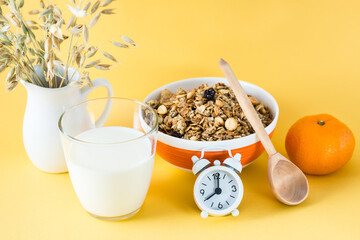 Healthy hearty breakfast. Baked granola in bowl, glass of milk, orange, wooden spoon and alarm...