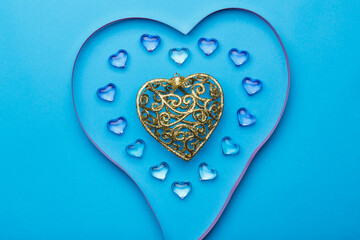 On a blue background, there is a heart made of transparent pebbles in the shape of hearts in the middle, a gold heart and a ribbon for decoration.
