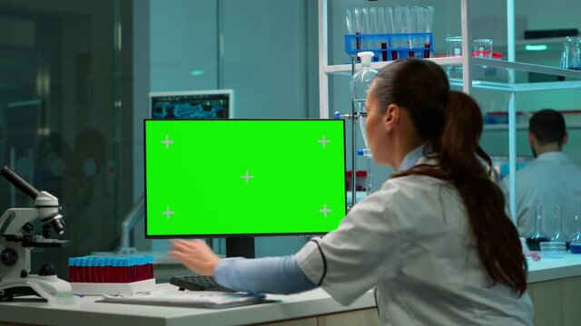 Scientist analysing blood sample and typing at computer with green screen, chroma key display. In background man lab researcher discussing with doctor about vaccine developent.
