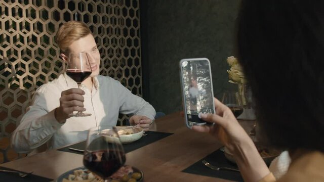 Over-the-shoulder medium shot of woman taking shot of her boyfriend sitting with glass of red wine at table in restaurant on smartphone