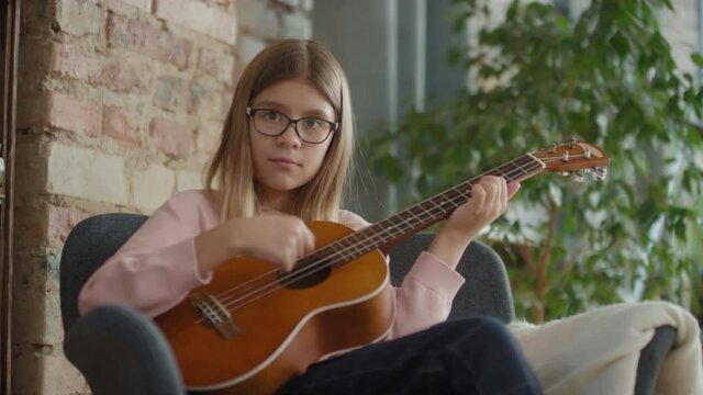Medium waist-up footage of talented girl learning how to play ukulele sitting at home watching online lessons