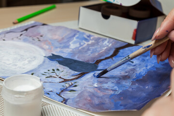 A close-up of female hands over a painted picture holding a brush and a cover for watercolors on which the colors are mixed next to the eraser and a simple pencil. A hobby of art.
