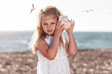 Little caucasian blond girl holding seashell and listening to the sea