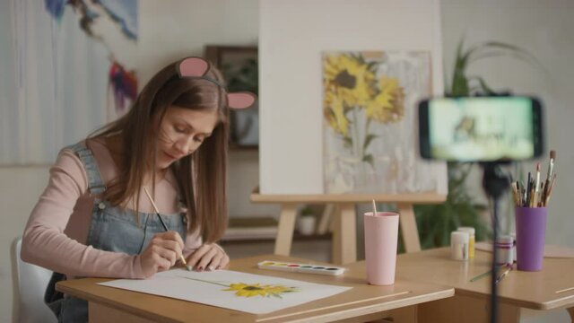 Medium shot of happy woman with mouse make-up and ears working as kids drawing teacher filming online lesson on smartphone sitting at home during lockdown