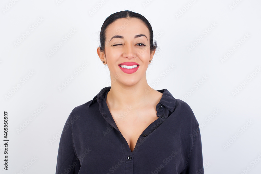 Sticker young beautiful Arab woman wearing gray dress against white studio background blinking eyes with pleasure having happy expression. Facial expressions and people emotions concept.