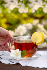 Female hands serve a glass cup with tea, lemon, embroidered napkin, jasmine branches on a wooden table. Outdoor, picnic, brunch.
