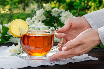 Female hands serve a glass cup with tea, lemon, embroidered napkin, jasmine branches on a wooden...