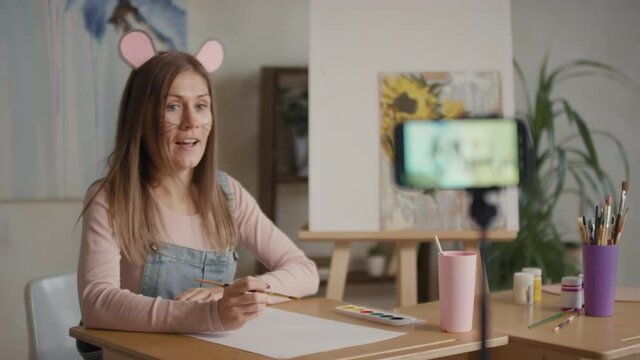 Medium shot of cheerful young female drawing teacher wearing mouse make-up and ears recording art lesson for kids with painted pictures in background