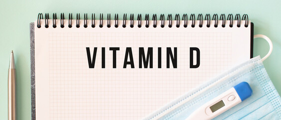 A medical mask and a thermometer are on the notepad. VITAMIN D text in a notebook.