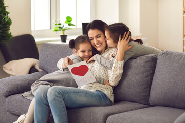 Obraz premium Happy young mother woman sitting at home on sofa and hugging two small beautiful kissing her daughters girls with room interior background. Mothers day, happy family and motherhood concept