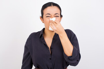 Young ill beautiful Arab woman wearing gray dress against white studio background sneezing her nose on a paper tissue feeling sick. Woman suffering from flu symptoms.
