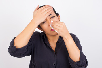Young ill beautiful Arab woman wearing gray dress against white studio background sneezing her nose on a paper tissue feeling sick. Woman suffering from flu symptoms.
