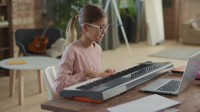 Medium shot of happy little girl having online music lesson via laptop at home learning to play keyboard