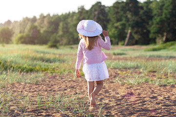 Backwards view of little girl in dress and white straw hat walking barefoot on sand at the sunset beach