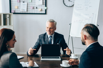 Smiling investor pointing at laptop with blank screen near business people with coffee on blurred foreground