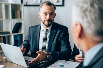 Businessman pointing at laptop near papers and investor on blurred foreground in office