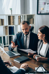 Businessman holding paper and pointing at investor on blurred foreground near laptop and colleague...