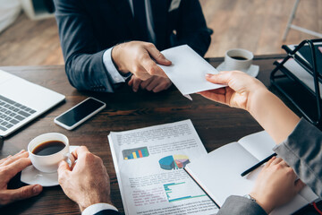 Cropped view of investor giving envelope to businesswoman near documents and cups of coffee on table