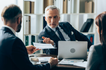 Mature investor pointing at businessman with document on blurred foreground near papers and laptop