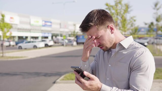A young Caucasian man works on a smartphone and acts frustrated in an urban area - a shopping center and a parking lot in the blurry background