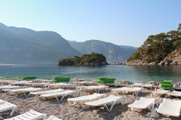 Fototapeta na wymiar Empty sun loungers and umbrellas facing turquoise water and Mountains by the beach on sunny day at blue lagoon of Oludeniz Fethiye Turkey