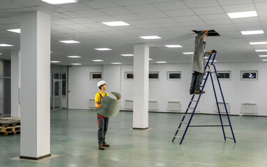 Workers on a ladder check the electrical wires on the ceiling. Workers in overalls check the correctness of the wiring under the ceiling in the office space. Check with the plan.