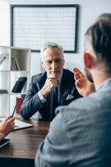 Investor in formal wear looking at businessman on blurred foreground near laptop in office