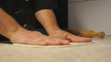 Obraz na płótnie Canvas Male arms of cook forming pastry on a wooden surface at restaurant. Hands of chef shaping floured dough for pizza on a kitchen table at cuisine. Concept of preparing food. Close up Slow motion