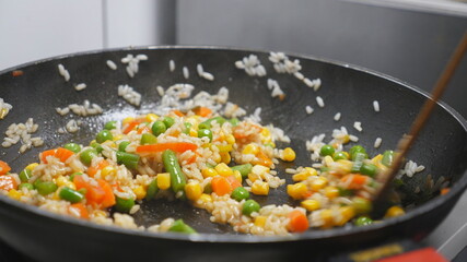 Male arm of chef stirring rice with mixed vegetables using a spatula on frying pan at cooker. Male hand of cook roasting risotto with corn, peas, asparagus, pepper and carrot at skillet. Close up