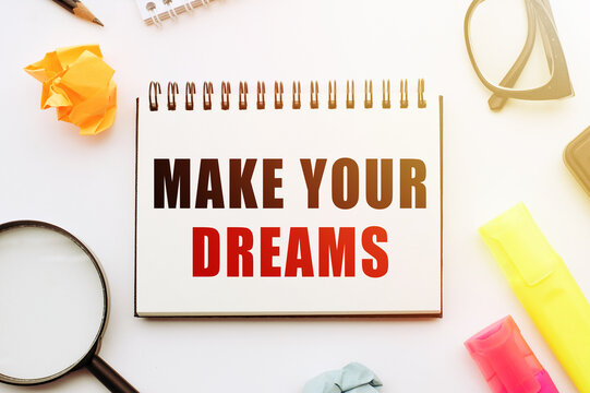 Text MAKE YOUR DREAMS in notebook on white table with office tools.
