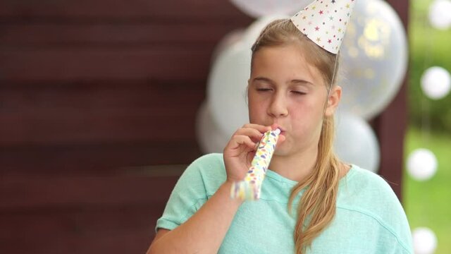 A teenage girl in a birthday hat blows a pipe and laughs. Happy childhood, children's birthday party outdoor