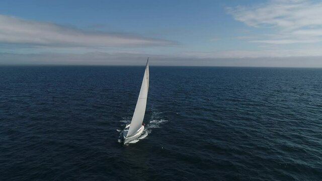 Aerial view. A white sailboat is moving fast on the waves of the sea. A boat in full sailing gear floats on camera. Close hauled sailing in ocean. Yacht in windy and sunny day. Drone shot.