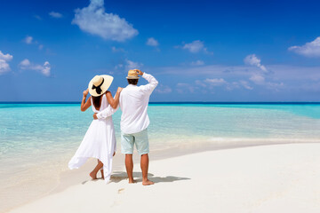 A beautiful tourist couple in white summer clothes and hats stands on a tropical beach and gazes at the turquoise ocean during their vacation time