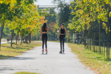 Rearview of two young fit women on roller skates riding outdoors on urban street in the park.