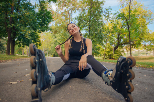 Astonishing young woman with hair braids posing in  roller skates while sitting on a road in the park
